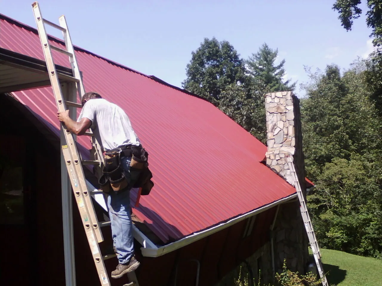 Roofer on a ladder beside a red metal roof and stone chimney — Soffit & Fascia Repair by Roofing Brothers in Valley Cottage
